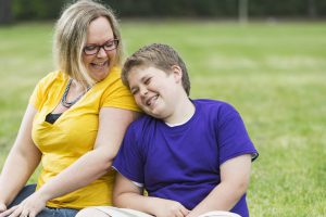 Mother and preteen son sitting together on the grass in the park, laughing. The boy is leaning his head against his mom's shoulder and she is looking down at him. They are wearing casual clothing, jeans, shorts, and t-shirts. They are ordinary people, heavyset, happy and confident. The woman is wearing eyeglasses.